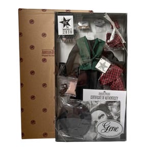 May include: A boxed Ashton-Drake Gene doll set featuring a dark green vest, brown long-sleeved shirt, and red plaid skirt. The package includes a black hat, small accessories in plastic bags, and a Certificate of Authenticity with the text Gene.