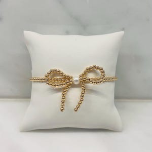 May include: Gold beaded bracelet with a bow design. The bow is made of small gold beads and features a single pearl in the center. The bracelet is displayed on a white pillow.