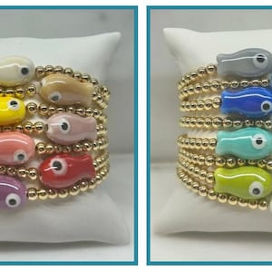 Puede incluir: Un conjunto de ocho pulseras de cuentas doradas con abalorios de peces de colores. Los abalorios de peces tienen un diseño de ojo malvado. Las pulseras están apiladas unas sobre otras.