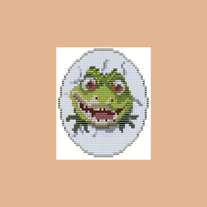 Könnte beinhalten: Ein Kreuzstich-Design einer grünen Froschfigur, die aus einem zerbrochenen weißen Ei kommt. Der Frosch hat große orangefarbene Augen und ein breites, zahnreiches Grinsen. Der Hintergrund ist hellbraun.