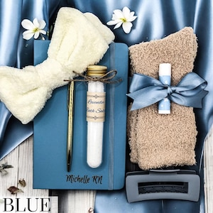 May include: A blue journal with the name "Michelle RN" engraved on the cover. The journal is surrounded by a white fluffy bow, a small glass tube filled with bath salts, a gold pen, and a brown fuzzy hand towel with a blue ribbon.