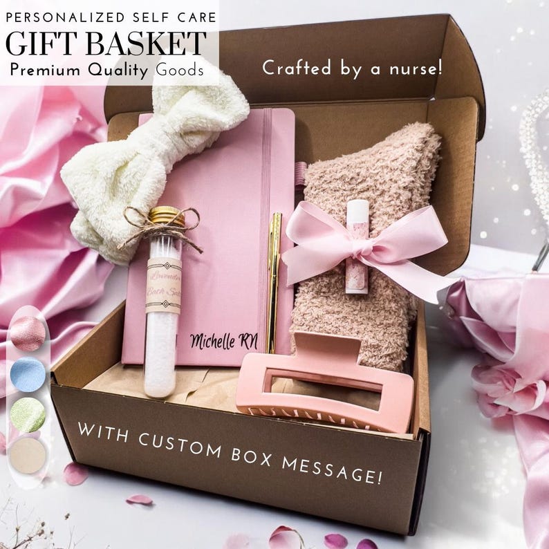 May include: A brown gift box with a pink and white bow, a pink journal with a gold pen, a pink headband, a small glass bottle with a white label, a pink fuzzy towel, a pink lip balm, and a pink hair clip. The box has the text "Personalized Self Care Gift Basket Premium Quality Goods Crafted by a nurse!" and "With Custom Box Message!" on it.