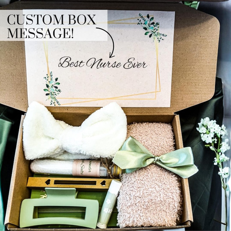 May include: A brown cardboard box with a green and gold floral design on the lid. The lid is open and the box is filled with various items, including a white headband, a bottle of bath salts, a green hair clip, a lip balm, and a pink fuzzy blanket. The lid of the box has the text "Custom Box Message!" and "Best Nurse Ever" written on it.