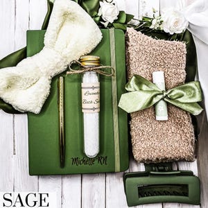 May include: A green journal with the name "Michelle RM" engraved on the cover, a white fluffy bow, a small glass bottle with a label that says "Lavender Bath Salts", a green hair clip, a brown towel with a green ribbon, and a lip balm.
