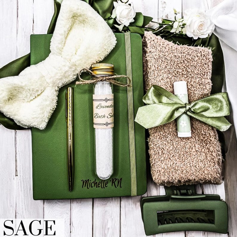 May include: A green journal with the name "Michelle RM" engraved on the cover, a white fluffy bow, a small glass bottle with a label that says "Lavender Bath Salts", a green hair clip, a brown towel with a green ribbon, and a lip balm.