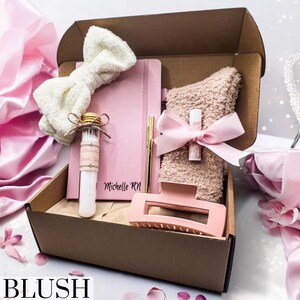 May include: A brown cardboard gift box with a pink bow and a pink notebook with the text "Michelle RN" on the cover. The box also contains a pink fluffy towel, a small glass bottle with a white label, a pink hair clip, and a gold pen.