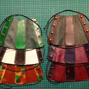 Könnte beinhalten: Vier Leder-Handgelenksmanschetten mit dekorativen Nähten und Messingakzenten. Die Manschetten sind in verschiedenen Farben übereinander gelegt, darunter Grün, Braun, Lila und Rot.