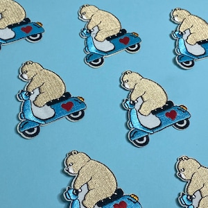 May include: Ten embroidered iron-on patches featuring a cartoon bear riding a blue scooter with a red heart. The patches are perfect for adding a whimsical touch to clothing, bags, or other fabric items.