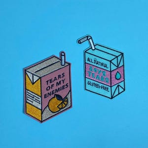 Op de afbeelding: Twee geborduurde strijkbare patches in de vorm van sappakken. Eén is roze en geel met de tekst "Tears of my enemies" en een schijfje sinaasappel. De andere is blauw en roze met de tekst "All Natural Boys Tears Gluten-Free".
