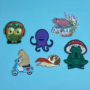 May include: Set of seven embroidered iron-on patches featuring various animals and designs. The patches include a green owl wearing headphones, a purple octopus, a frog meditating on a mushroom, a frog in a teacup, a bear riding a scooter, a pug wearing a cape, and a green frog meditating on a mushroom.
