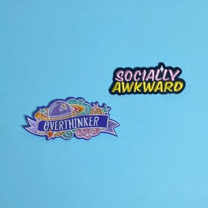 Pode incluir: Dois patches bordados para passar a ferro. Um patch é roxo, rosa e amarelo com um planeta e o texto "Overthinker". O outro patch é preto com texto amarelo que diz "Socially Awkward".