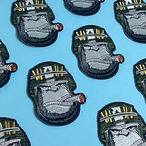 Può includere: Un set di nove toppe ricamate con un gorilla dei cartoni animati che indossa un elmetto verde dell'esercito con munizioni dorate e tiene un sigaro. Il gorilla ha un'espressione seria e guarda a destra.