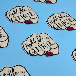 May include: A set of six iron-on patches featuring a fist with the text "Fight Like a Girl" in black cursive lettering. The fist is beige with red nail polish.