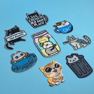 May include: Nine embroidered iron-on patches featuring cute cats in various poses and with funny sayings. The patches include a cat in a coffee cup saying "Coffee is my safe place", a cat in a bin, a cat holding a knife, a cat in a jar with other cats, a cat in a cup saying "Cat Puccino", a cat with the saying "Your poop smells like flowers", and a cat in a cup saying "OK".