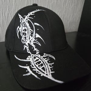 May include: Black baseball cap featuring a white tribal design. The cap has a curved peak and a button on top. The white design is symmetrical and abstract, with sharp lines and curves.