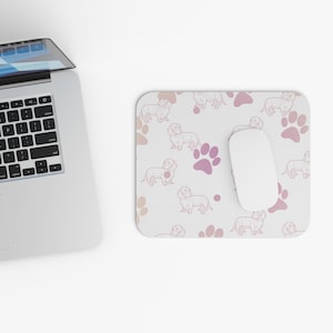 May include: A rectangular mousepad with a white background, featuring a repeating pattern of dachshund dogs and paw prints in shades of pink and peach. A white computer mouse rests on the pad, next to a laptop.