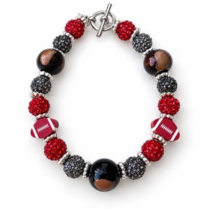 May include: A beaded bracelet featuring red and black rhinestone-encrusted spheres, smooth black beads, and two red football-shaped charms with white laces. The bracelet is secured with a silver-toned toggle clasp and accented with small silver spacers.