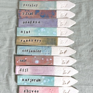 May include: A selection of ceramic herb garden markers. Each marker features a coloured top section and a white, pointed base. The names of the herbs are: thyme, parsley, basil, oregano, mint, rosemary, coriander, sage, dill, marjoram, chives, and tarragon.