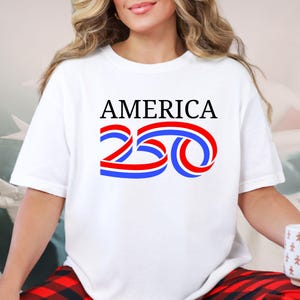 Może przedstawiać: Biała koszulka z napisem "AMERICA" w czarnych literach nad stylizowanym wzorem "250" w kolorze czerwonym i niebieskim. Koszulka ma swobodny styl.