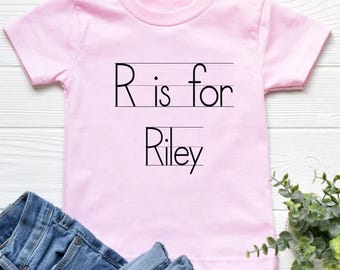 May include: A light pink t-shirt with the text "R is for Riley" in black. The shirt is short-sleeved and has a round neckline. Blue denim jeans are partially visible below the shirt.