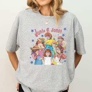 Op de afbeelding: Grijs T-shirt met een grafische print van de personages uit het boek "Junie B. Jones" van Barbara Park. De afbeelding toont Junie B. Jones en haar vrienden.