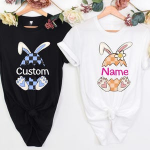 Może przedstawiać: Dwie białe koszulki z królikiem wyłaniającym się z jajka wielkanocnego. Jedna koszulka ma napis "Custom", a druga "Name".