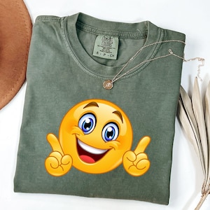 May include: An olive green t-shirt with a large, smiling emoji graphic. The emoji has a yellow face, blue eyes, and raised index fingers. A silver necklace with a small pendant rests on the shirt. The shirt is labeled "Comfort Colors" and is size small.