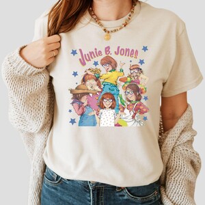 Op de afbeelding: Een beige T-shirt met een grafische print van de personages uit de boekenserie "Junie B. Jones". De grafiek toont de personages Junie B. Jones, haar vrienden en de tekst "Junie B. Jones!" in roze en paarse letters.