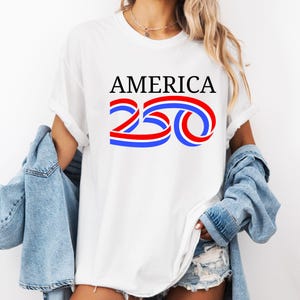 Może przedstawiać: Biała koszulka z napisem "AMERICA" w czarnym kolorze nad czerwono-niebieskim wzorem "250". Koszulka jest noszona z dżinsową kurtką.