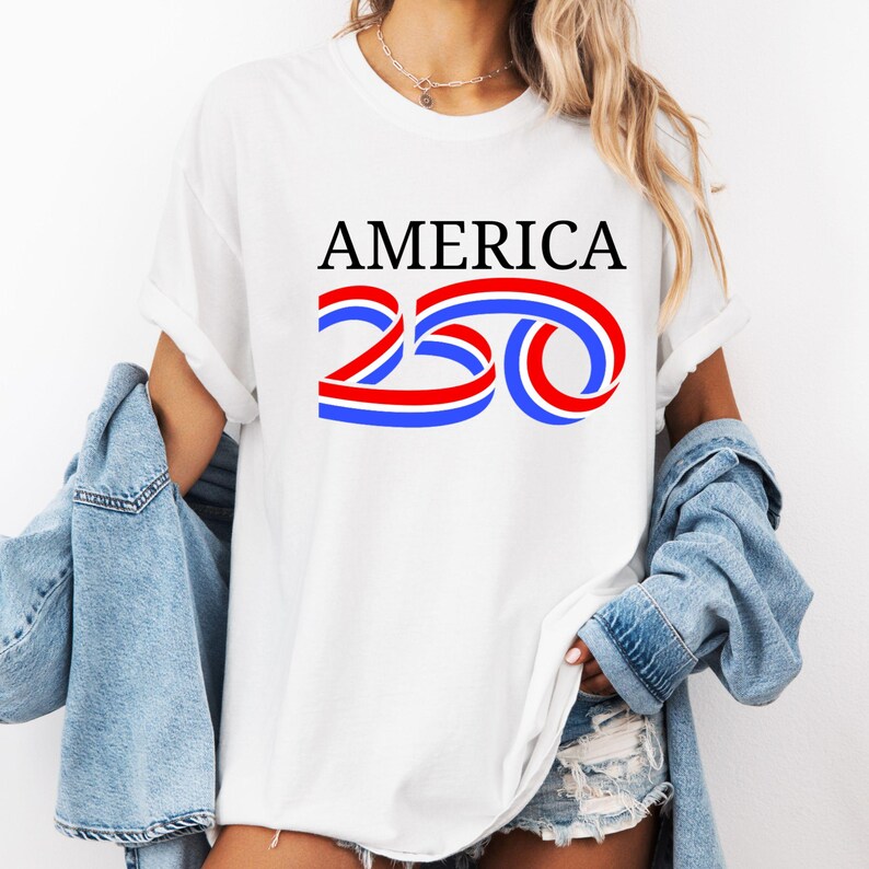Może przedstawiać: Biała koszulka z napisem "AMERICA" w czarnym kolorze nad czerwono-niebieskim wzorem "250". Koszulka jest noszona z dżinsową kurtką.