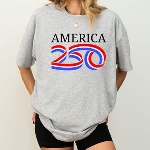 Może przedstawiać: Szara koszulka z napisem "AMERICA" w kolorze czarnym oraz liczbą "250" w kolorze czerwonym, białym i niebieskim. Koszulka jest w stylu casual, odpowiednia do codziennego użytku.