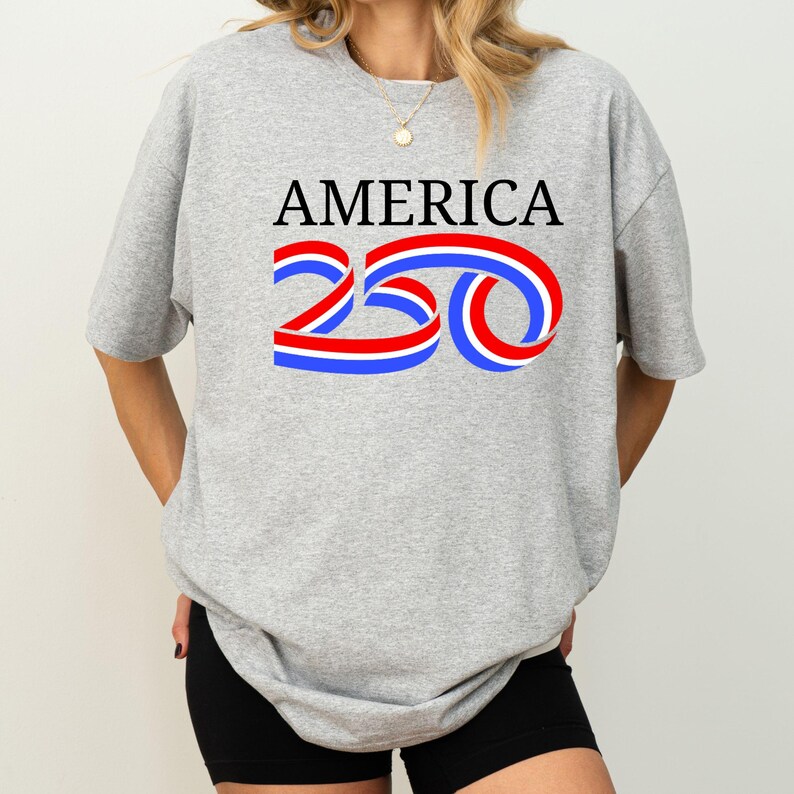 Może przedstawiać: Szara koszulka z napisem "AMERICA" w kolorze czarnym oraz liczbą "250" w kolorze czerwonym, białym i niebieskim. Koszulka jest w stylu casual, odpowiednia do codziennego użytku.