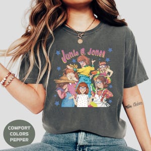 Op de afbeelding: Een donkergrijs T-shirt met een afbeelding van de personages uit de Junie B. Jones-boekenreeks. De afbeelding toont de personages Junie B. Jones, haar vrienden en haar lerares. De tekst "Junie B. Jones" is in roze en paarse letters op het shirt gedrukt.