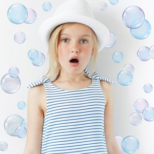 May include: A young girl wearing a white hat and a blue and white striped dress looks surprised as she stands in front of a white wall with blue and purple bubble stickers.