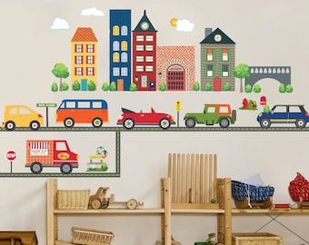 May include: A colorful city scene wall decal with buildings, cars, and a road. The decal is applied to a white wall in a playroom with wooden shelves and toy cars.