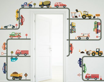 May include: A set of colorful vehicle wall decals with roads and traffic signs. The decals include a police car, fire truck, ambulance, construction vehicles, and other cars. The decals are arranged on a white wall with a door in the middle.
