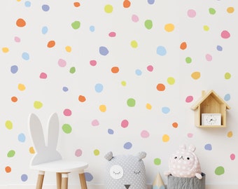 May include: A white wall with colorful polka dots in various shades of pink, blue, green, yellow, and orange. The wall is decorated with a white bunny chair, a gray pillow with a white face, and a pink bunny toy in a gray basket.