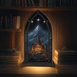 Op de afbeelding: Een decoratief glas-in-loodraam met een berg en een stad met verlichte gebouwen. Het raam is zwart omlijst en staat op een houten oppervlak. Boekenkasten vol boeken staan op de achtergrond.