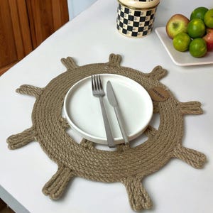 May include: A brown, rope-like placemat shaped like a ship's wheel with a white plate, fork, and knife on top. The placemat has a small tag that says "Handmade".
