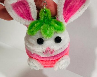 May include: A handmade bunny keychain crafted from white, pink, and green pipe cleaners. The bunny features large pink-lined ears, black bead eyes, and a small pink nose. The keychain is attached to a silver metal chain.