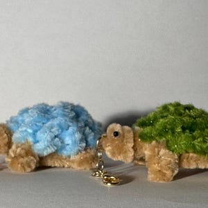 May include: Two plush turtle keychains. One has a light blue shell, the other a green shell. Both have tan bodies, black eyes, and gold-colored key rings. The turtles are made of soft, fuzzy material.