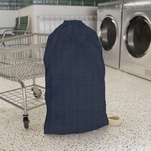 May include: A large, navy blue laundry bag with a plaid pattern. The bag is made of fabric and has a drawstring closure at the top. The bag is standing upright on a speckled floor.