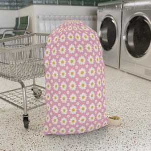 May include: A pink laundry bag with a daisy pattern. The bag is filled and upright, with a drawstring closure at the top. The daisies are white with yellow centers. A laundry cart is in the background.