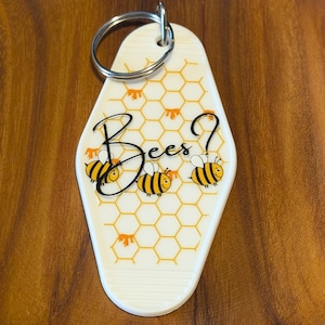 Può includere: Portachiavi bianco in stile motel con portachiavi argentato. Il portachiavi presenta un motivo a nido d'ape e tre api dei cartoni animati. La parola "Bees?" è scritta in corsivo nero.