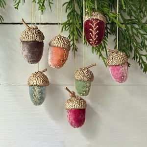 May include: A collection of seven hanging acorn ornaments featuring soft velvet bodies in shades of deep red, pink, orange, green, and brown. Each acorn has a textured gold cap and a delicate gold stem, with gold thread embroidery detailing.
