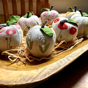 May include: A collection of round, fabric-covered decorative pumpkins in a wooden bowl. Each pumpkin is made of light beige linen fabric and features detailed embroidery of various flowers, including red poppies and white lily of the valley.