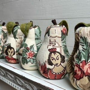May include: A row of five pear-shaped fabric ornaments with a green leaf stem. Each pear is covered in a different patterned fabric featuring floral and human figures. The fabric is a mix of green, white, red, and pink.