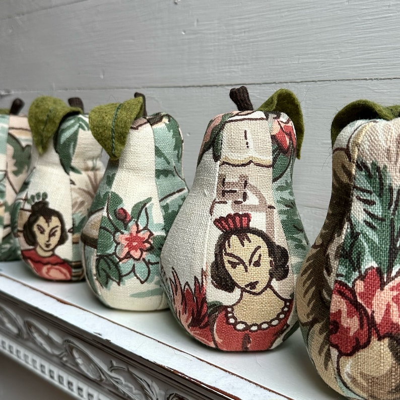 May include: A row of five pear-shaped fabric ornaments with a green leaf stem. Each pear is covered in a different patterned fabric featuring floral and human figures. The fabric is a mix of green, white, red, and pink.