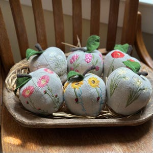 May include: Six gray fabric apple-shaped ornaments with embroidered floral designs, including daisies, sunflowers, and poppies. Each apple has a green leaf and a brown stem. They are arranged in a wooden bowl.