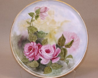 May include: A white porcelain plate with a gold rim features a hand-painted floral design. The plate depicts pink roses and green leaves against a light blue background.